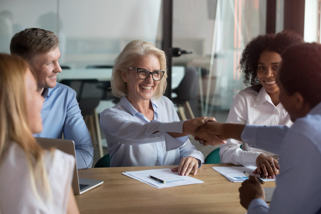 Middle aged woman executive manager company representative welcoming business partner while sitting in modern office boardroom or trainer shake hands with participant before starting education meeting
