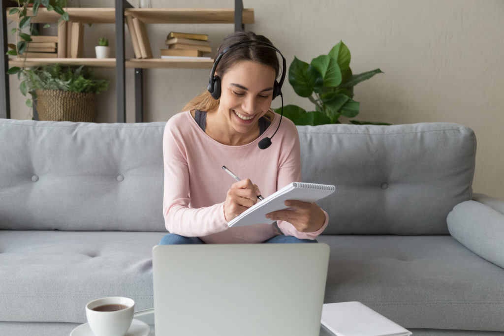 Happy woman sitting on sofa wearing headset with microphone learning English with teacher online, study improve knowledge using computer apps, holds pen noting in notepad feels satisfied concept image