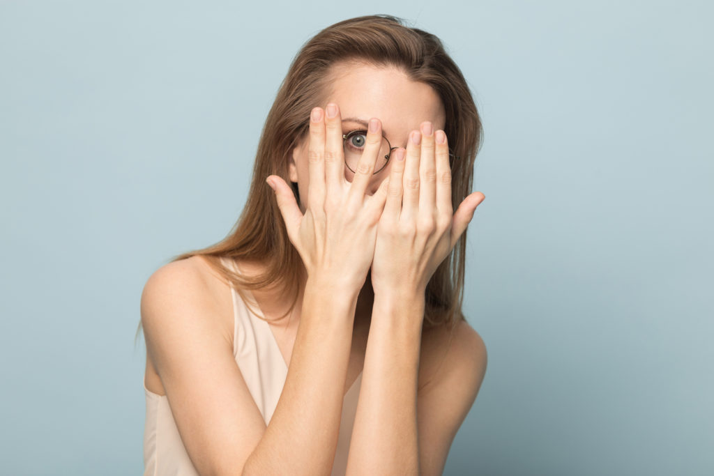 Scared pretty young woman in glasses hiding behind hands, peeping through fingers, curious funny female looking at camera, feeling afraid or shy, peeking, isolated on studio background