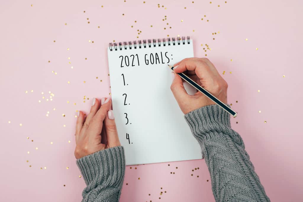 Woman's hand writing 2021 Goals in notebook decorated with Christmas decorations on the tricolor background. Top view, flat lay